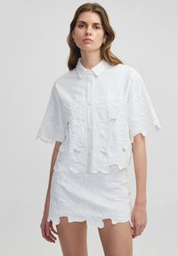 Camicia bianca a maniche corte, con ricamo di design floreale, collo strutturato e chiusura a bottoni; abbinata a una gonna coordinata con dettagli simili.