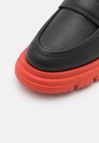 Mocassin en cuir noir avec une semelle en caoutchouc rouge épaisse et audacieuse, bout arrondi, présenté en gros plan sur fond blanc.