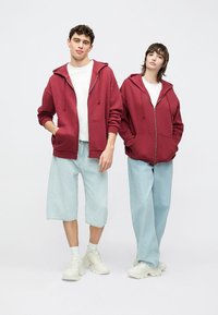 Két modell visel ugyanolyan borvörös cipzáras kapucnis pulóvert elülső zsebekkel, világoskék farmer rövidnadrággal és fehér tornacipővel párosítva.
