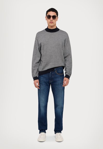 Maglione a righe grigie con colletto a coste nero, abbinato a jeans blu a gamba dritta e sneakers bianche. Occhiali da sole rotondi neri.