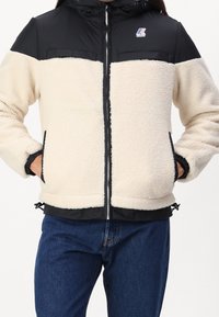 Valkoinen ja musta vetoketjullinen takki, jossa fleece-vartalo, nylon-hartiot, elastiset hihansuut, sivutaskut ja säädettävä helma. Logo rintakohdalla.