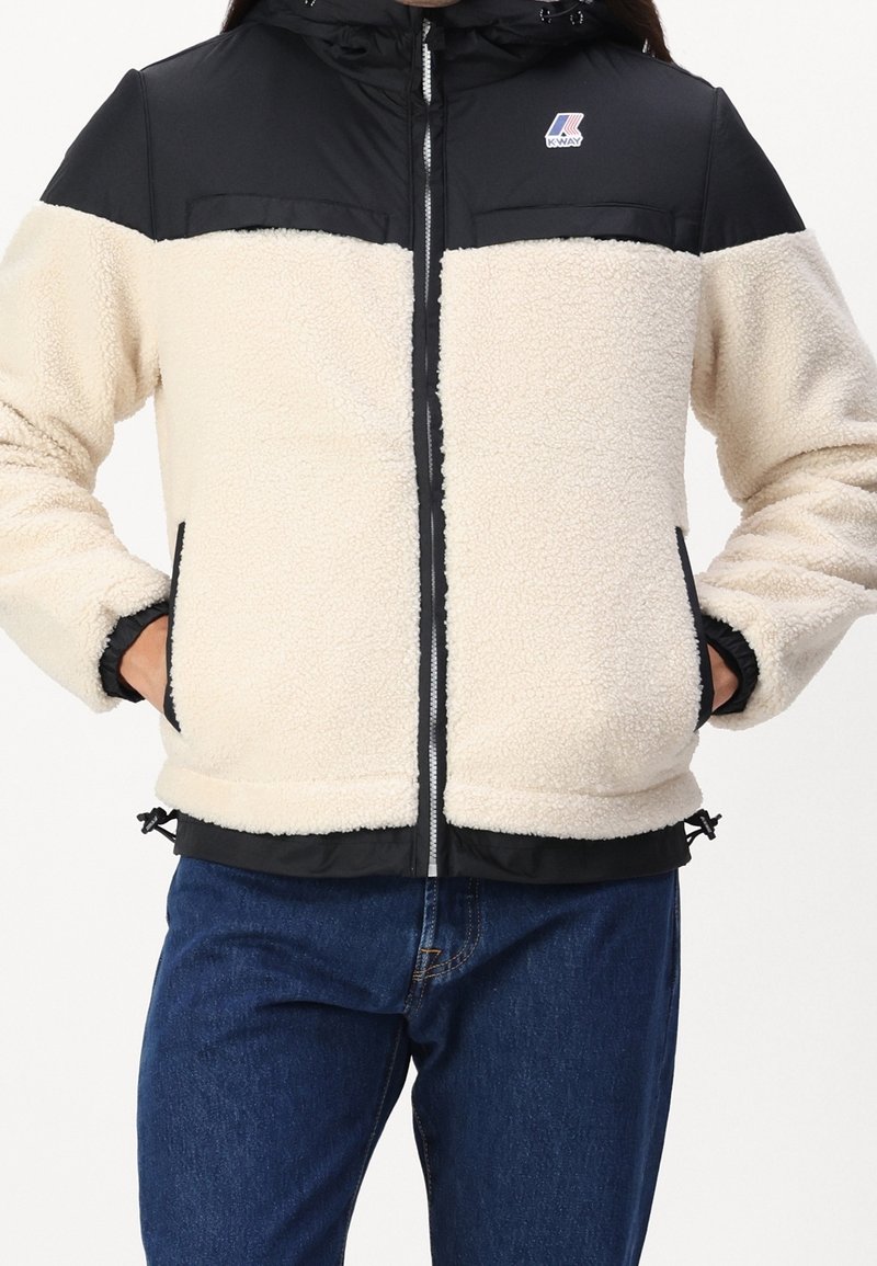 Valkoinen ja musta vetoketjullinen takki, jossa fleece-vartalo, nylon-hartiot, elastiset hihansuut, sivutaskut ja säädettävä helma. Logo rintakohdalla.