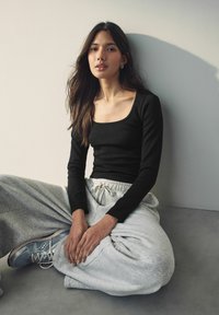 Zwarte ribbelige langemouwen top gecombineerd met grijze sweatpants. Model zit met handen op de knieën, draagt blauwe sneakers. Eenvoudige, casual outfit.
