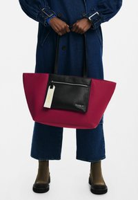 Personne portant un manteau en jean et un jean court, tenant un grand sac cabas bicolore bordeaux et noir avec une poche zippée à l'avant.
