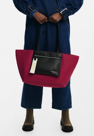 Persona con cappotto di denim e jeans corti che tiene una grande borsa tote bicolore bordeaux e nera con tasca frontale con cerniera.