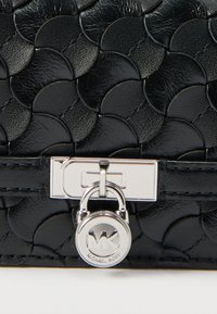 Juodos odos rankinė su tekstūruotu žvynelių raštu, sidabrinės spalvos furnitūra ir apvali užrakinimo detalė su prekės ženklo logotipu.