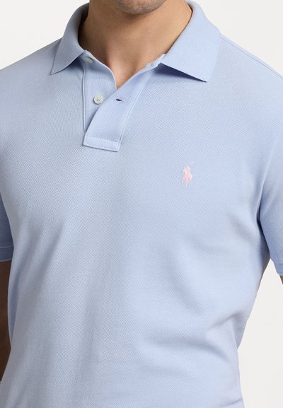 Gaiši zilas polo krekls no teksturēta auduma ar divu pogu apkakli. Uz kreisā krūts ir mazs rozā logotips.