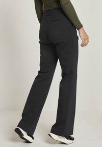 Person som bär svarta vida byxor och vita sneakers, står med ena foten något höjd, mot en enkel ljus bakgrund.