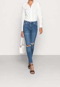 Light blue skinny jeans with distressed knees, paired with a fitted white button-up shirt and black strappy heels, carrying a white handbag.