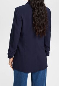 Kvinna med långt vågigt mörkt hår, iklädd en marinblå blazer och blå jeans, sedd bakifrån mot en enkel vit bakgrund.