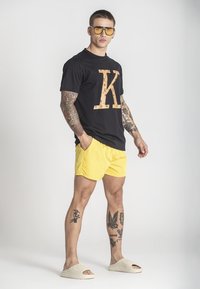 Černé tričko s velkým oranžovým logem 'K', žluté plavky, lehké nazouvací boty a brýle se žlutým nádechem; viditelné tetování na rukou a nohách.