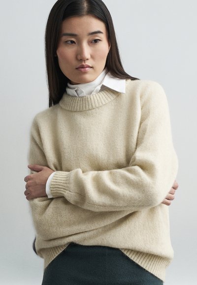 Pull en crème texturé avec un col et des poignets côtelés, porté par-dessus une chemise blanche à col. Coupe loose et fentes latérales subtiles.