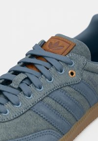 Κοντινό πλάνο ενός μπλε δερμάτινου και σουέτ αθλητικού παπουτσιού Adidas με ανοιχτό μπλε κορδόνια και καφέ δερμάτινο λογότυπο στη γλώσσα.