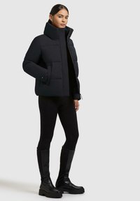Donna che indossa un giubbotto imbottito nero, leggings neri e stivali neri fino al ginocchio, in piedi su uno sfondo neutro.