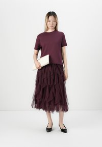 Woman wearing a burgundy short-sleeve top and tiered tulle midi skirt, holding a beige clutch and wearing black slingback heels.