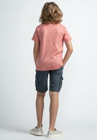 Lyhythihainen, korallinpunainen t-paita, jossa on teksturoitu kuvio, yhdistettynä harmaisiin cargo-shortsihin, joissa on sivutaskut ja vaaleanbeigeet slip-on-kengät.