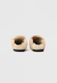 Pantofole beige foderate in pelliccia, con una texture morbida, design aperto sul retro e suola in gomma nera. Profilo piatto adatto per l'uso indoor.