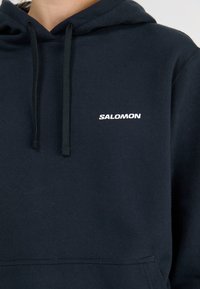 Tumesinine kapuutsiga sviiter, mis on valmistatud puuvilla segust, koos esiseguga. Ees on valge "SALOMON" logo ja kapuutsil on nöörid.