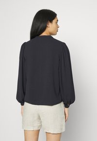 Blusa azul-marinho com mangas bufantes, feita de um tecido leve, apresentando um decote redondo e detalhes franzidos nas costuras dos ombros.