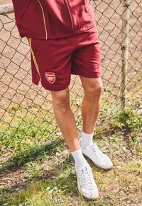 Bordeaux atletiske shorts med gule striper, med Arsenal-logo. Kombinert med hvite sokker og sneakers. Gress og gjerde i bakgrunnen.