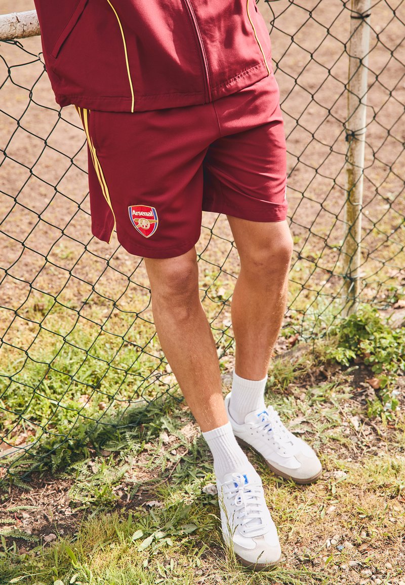 Bordeaux atletiske shorts med gule striper, med Arsenal-logo. Kombinert med hvite sokker og sneakers. Gress og gjerde i bakgrunnen.