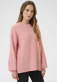 Hellrosa, locker sitzender Pullover mit Rundhalsausschnitt und langen Ärmeln. Weiche Textur und gerippte Bündchen. Kombiniert mit dunklen Hosen.