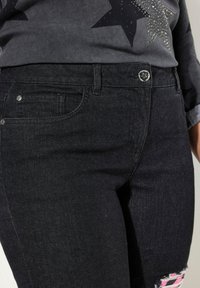 Jeans in denim nero con un design aderente, tasche frontali, bottone argentato e dettagli strappati con accenti rosa sul ginocchio.