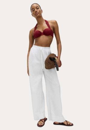 Kobieta stoi ubrana w bordowy bikini z wiązaniem na szyi, białe, szerokie spodnie, brązową plecioną kopertówkę i ciemnobrązowe płaskie sandały na tle o jednolitym kolorze.