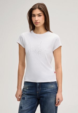 Jonge vrouw draagt een witte aansluitende T-shirt met tekst 'Joop! Jeans' van strass-steentjes en beschadigde blauwe spijkerbroek tegen een effen achtergrond.