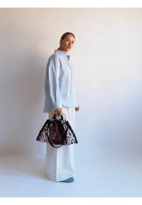 Chemise longue en lin bleu clair à manches longues, pantalon blanc à jambes larges et sac à main imprimé léopard avec garniture noire, tenu dans une main.