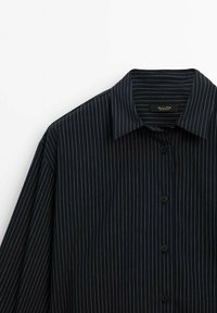 Chemise noire à manches longues avec fines rayures verticales blanches, devant boutonné et col pointu sur fond blanc.