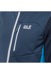 Mörkblå jacka med en slät yta, med en vit dragkedja och logotyp. Inkluderar en ficka med dragkedja och en kontrasterande blå dragning.