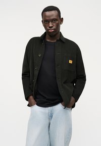 Jachetă cămașă din catifea de culoare verde închis cu un buzunar la piept și etichetă galbenă, purtată peste un tricou negru și blugi din denim de culoare albastru deschis.