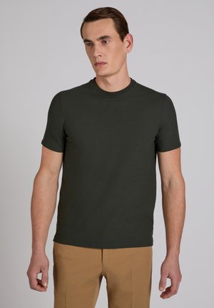 Uomo che indossa una maglietta verde scuro con scollo rotondo e pantaloni marrone chiaro, in piedi con le braccia rilassate contro uno sfondo grigio uniforme.