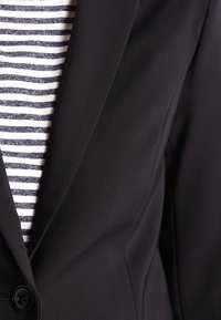Nahaufnahme eines schwarzen Blazers über einem weiß-blauen horizontal gestreiften Hemd, das den Revers und den Knopf des Blazers hervorhebt.