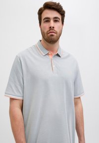 Světle šedé polo tričko s límcem v oranžovo-bílých pruzích. Má tři knoflíky u krku a jemnou texturu. Bez vzorů.