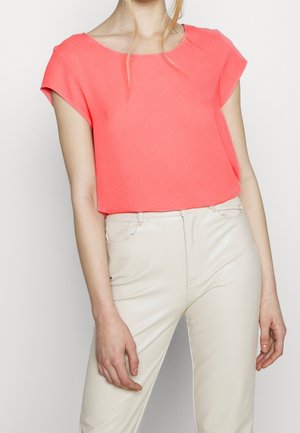 Femme portant un chemisier corail à manches courtes, rentré dans un pantalon taille haute couleur ivoire, debout devant un fond uni.