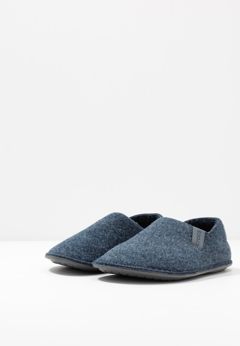 Pantuflas de fieltro azul oscuro con una textura suave, forma de punta afilada y una discreta etiqueta con logo. Suela de goma negra para agarre y durabilidad.