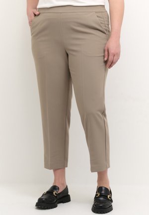 Beige, gekürzte Hose mit Seitentaschen, getragen mit schwarzen Loafers, an einer stehenden Person vor einem einfarbigen Hintergrund.
