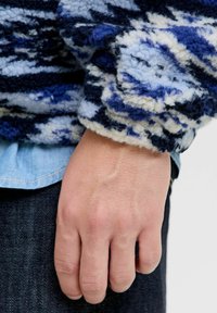 Przytulny, fakturowany sweter w odcieniach niebieskiego i białego, z puchatym, abstrakcyjnym wzorem. Rękawy typu mankiet i założony na dżinsową koszulę.