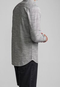 Camicia grigia a maniche lunghe con tessuto texturizzato, orlo arrotondato e polsini con bottoni, indossata sopra pantaloni corti scuri. Mostra il design della schiena e delle spalle.