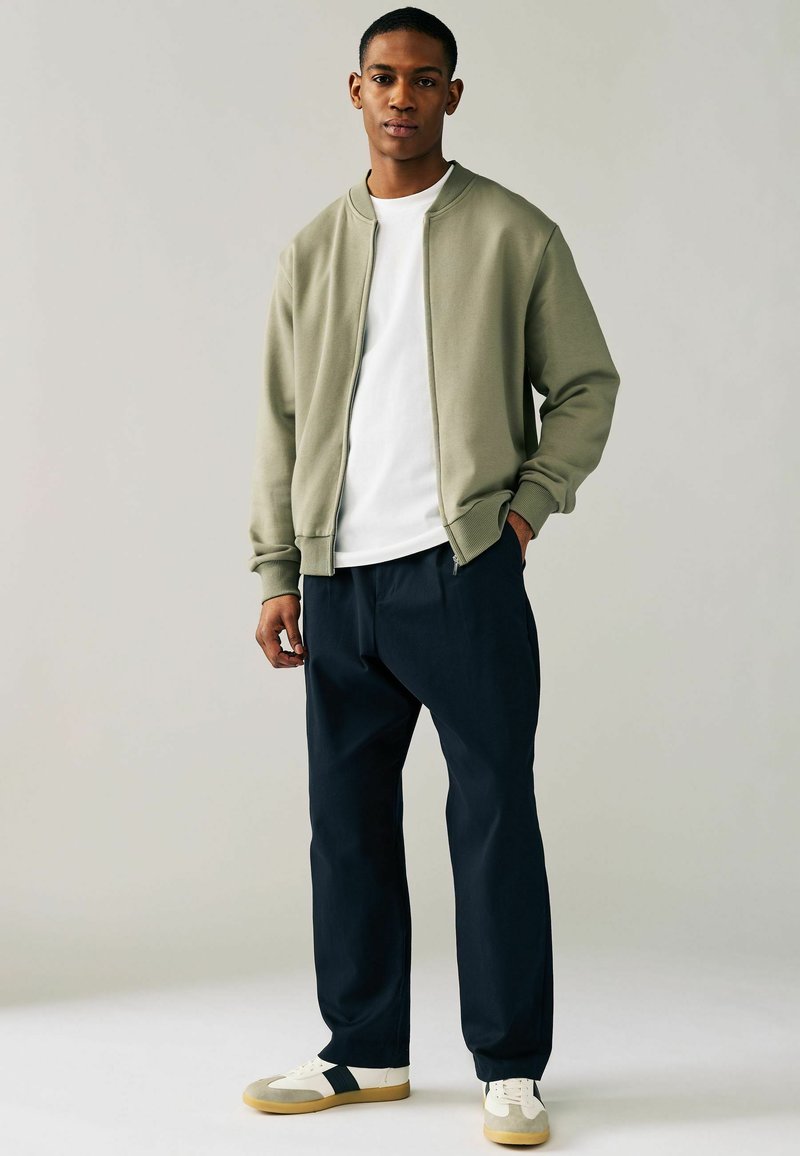 Chaqueta bomber verde sobre una camiseta blanca, combinada con pantalones navy y zapatillas blancas con detalles en gris. Diseño simple y casual.