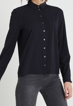 Femme portant une chemise noire à manches longues avec de petits pois blancs et un jean ajusté gris foncé.