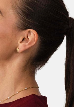 Frau mit dunklem, zurückgebundenem Haar, die einen kleinen goldenen drachenförmigen Ohrring und eine goldene Halskette mit abgesetzten weißen Perlen trägt, vor weißem Hintergrund.