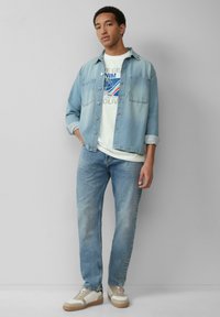 Jasnoniebieska kurtka dżinsowa na białym t-shircie z grafiką, w połączeniu z jasnoniebieskimi dżinsami i beżowymi sneakersami z brązowymi podeszwami. Prosty design, swobodny fason.