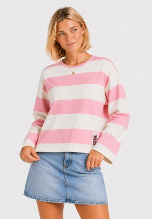 Kobieta z krótkimi blond włosami nosi różowo-białą, w paski, bluzkę z długim rękawem oraz niebieską dżinsową spódnicę, patrzy na bok z lekkim uśmiechem.