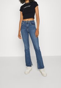 Sort cropped T-shirt med "Levi's" logo, parret med blå flagrende jeans med sideslids. Modellens fodtøj er hvide sneakers.