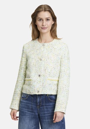 Giacca testurizzata di un leggero giallo con accenti verdi e blu, colletto rotondo, chiusura a bottoni e due tasche frontali. Indossata sopra dei jeans.