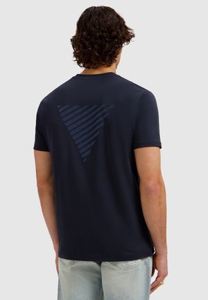Mann mit lockigem dunklem Haar, der ein marineblaues T-Shirt mit einem dunklen, gestreiften Dreiecksmuster auf der Rückseite trägt, kombiniert mit hellblauen Jeans.