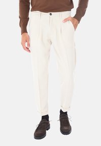 Pantaloni beige plissettati con vestibilità rilassata, dotati di tasche laterali, chiusura con un solo bottone e orli a risvolto, abbinati a scarpe marroni scure.